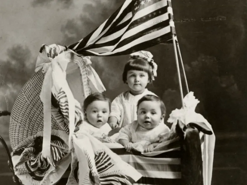 Three children in a wicker baby carriage with an American flag.
